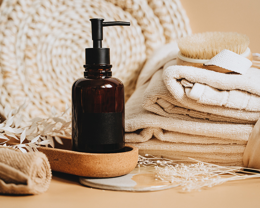Domáci šampón na vlasy pre začiatočníkov v hnedom skle s pumpičkou sotjí na stole v zátiší s uterákmi, kefou a inými umývacími prostriedkami.